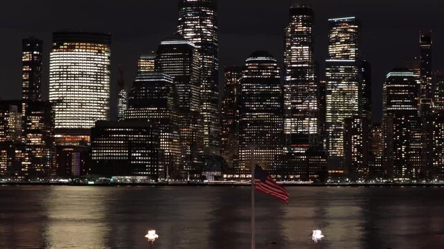 Aerial night view of New York City skyline with illuminated skyscrapers and reflections on river, with an American flag in foreground during winter snowfall. Patriotic urban scene in USA. Wide shot.