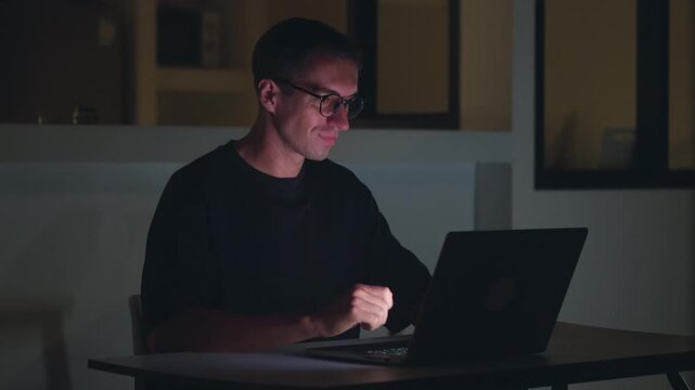 Man working late at night on laptop, typing code with focused expression and low light. Night work reflects overtime, dedication, productivity, deadlines and intense modern workflow