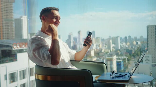 Happy businessman celebrates online win using smartphone while sitting by window with city view. Man shows joy after winning money on mobile app, digital success, fintech, online casino concept.