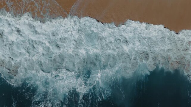 Cinematic aerial slow motion of ocean waves crashing onto sandy beach. Powerful sea foam and blue water form abstract coastal texture ideal for travel visuals nature backgrounds cinematic footage