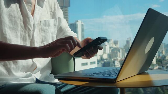 Businessman using smartphone near laptop by window with city skyline. Remote work professional checking phone and laptop in modern hotel office, business travel remote work lifestyle, digital nomad 