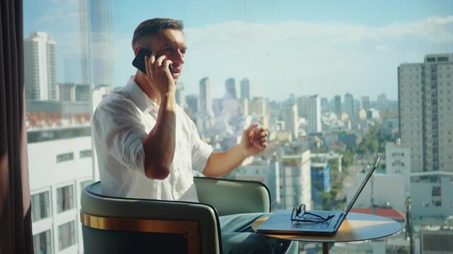 Confident businessman talking on smartphone while working on laptop near panoramic window with city skyline. Remote work and online business concept shows professional communication startup lifestyle.