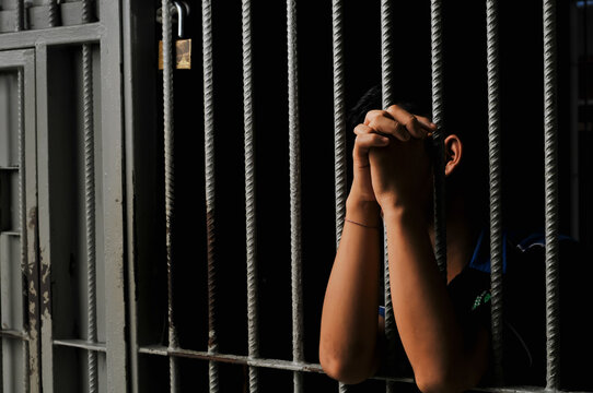 Close up of a prisoner hand gripping rusty iron bars behind a jail cell or detention center.
