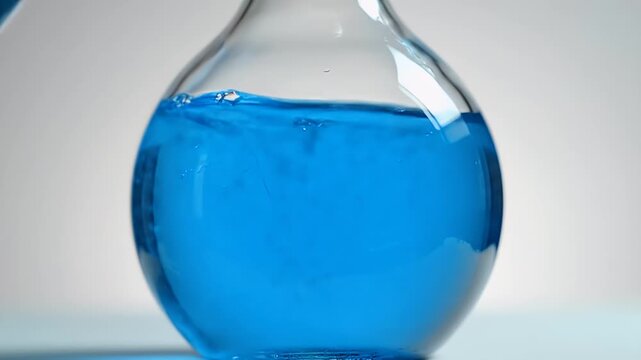 Close-up of a transparent glass round-bottom laboratory flask containing bright blue liquid on a soft neutral background