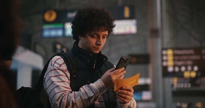 Young Man With Curly Hair, Wearing Striped Shirt and Navy Vest, Stands in Modern Airport Departure Hall, Uses Smartphone Scan Digital Ticket or Check In, Reviews Flight Details and Gate Information.