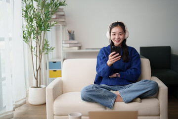 Fototapeta premium Happy young asian woman in casual attire enjoying leisure time, relaxing on a sofa at home, wearing headphones, and using a smartphone for music or entertainment