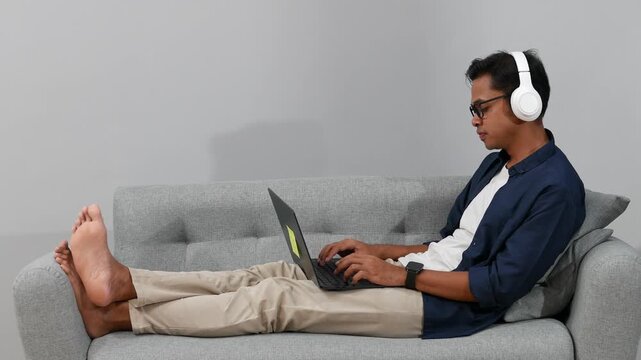 A side view of an Asian young man lying comfortably on a grey fabric sofa, wearing white headphones and using a laptop on lap