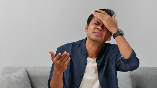 Confused young asian man sitting on sofa at home with asking hand gesture for choice, option and question
