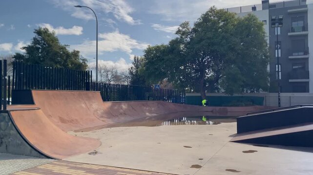 Urban skatepark bowl filled with rainwater, reflecting a person in a high visibility vest near a green tree under a cloudy sky