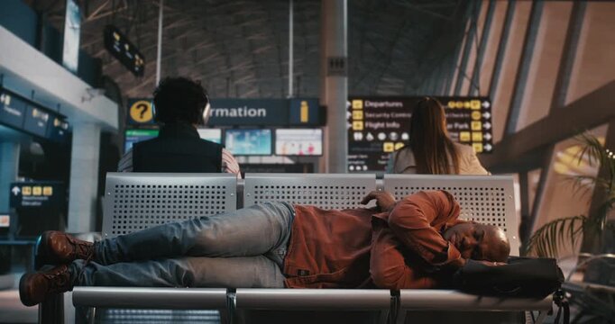 After Lengthy Flight Delay, Black Man Who Exhausted Falls Asleep While Lying Across Row of Metal Chairs in Departure Lounge. Males Backpack Acts Makeshift Pillow. Exhaustion of International Transit.