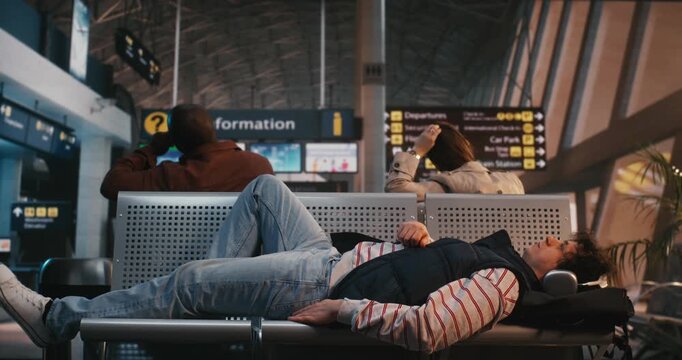 Weary Traveler in Striped Shirt and Vest Tries to Find Comfortable Position on Airport Seating While Waiting For Flight. Guy Falls Asleep at Airport. Physical Toll of Travel, Frustration of Delays.