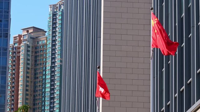 People's Republic of China and Hong Kong SAR flags wave against a backdrop of modern skyscrapers. Establishing shot of national identity and administrative sovereignty in a major Asian hub.