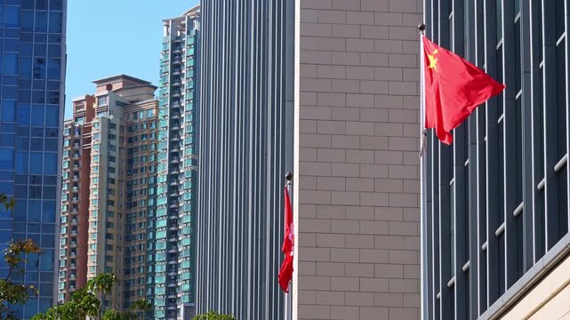 Chinese and Hong Kong flags fluttering on a sunny day in a dense commercial district. The shot illustrates the unique political landscape and urban architecture of the metropolis.