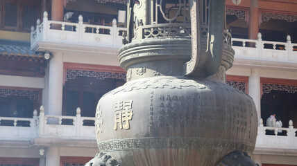 A bronze bell with intricate designs stands prominently in front of an Asian style building with traditional architecture © MakeitEZ-Img&VID
