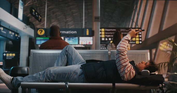 Young Man With Curly Hair, Wearing Large Headphones, Lies Across Row of Airport Chairs in Departure Lounge. Exhausted by Long Wait or Flight Delay, Male Holds Smartphone Above Him, Absorbed in Screen.