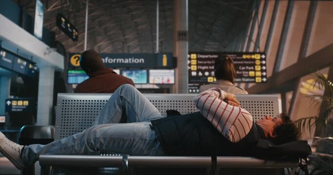 Exhausted Traveler Adjusts Position on Cold Metal Airport Chairs, Tucking Black Backpack Under Head For Comfort. After Long Wait in Terminal, Man Closes Eyes to Catch Some Sleep During Flight Delay.