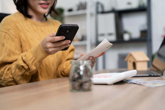 Asian woman using smartphone to pay bills or manage finances at home office desk with receipt, coins in jar, and calculator. Concept of mobile banking and financial planning.