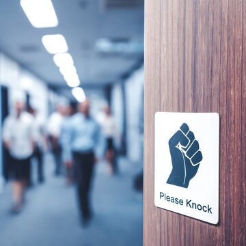 White please knock door sign featuring a black fist icon mounted on a professional wooden door with a busy corporate office hallway in the sharp background.