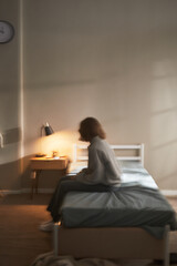 Vertical of Caucasian young adult woman sitting on bed in softly lit room, gazing downward with contemplative expression, hands resting on lap, conveying introspective emotion