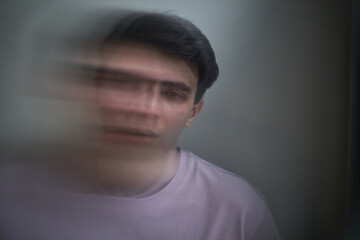 Portrait of young adult Caucasian man looking into camera with blurred double exposure effect suggesting complex emotion, short dark hair, neutral background, subtle facial expression