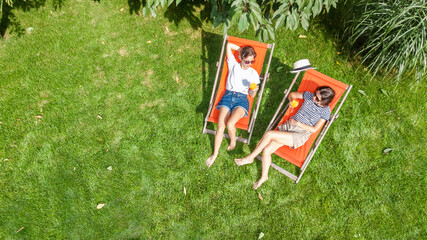 Young girls relax in summer garden in sunbed deckchairs on grass, women friends having drinks outdoors in green park lawn on weekend, aerial drone view 
