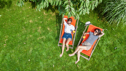Young girls relax in summer garden in sunbed deckchairs on grass, women friends having drinks outdoors in green park lawn on weekend, aerial drone view 