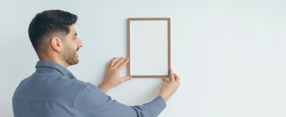 A man stands next to a wall, holding a blank frame with both hands. He is smiling as he prepares to hang the frame for decoration. The setting indicates a home improvement project during the daytime. © Prostock-studio