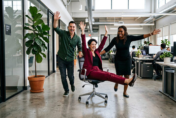 Fototapeta premium Three cheerful diverse business people enjoying ride on office chair in bright office.