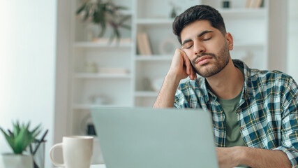 A young man sits at a desk with his hand on his face, appearing tired and distracted. He works on a...