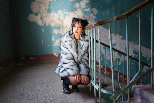 Woman in fur coat posing by stair railing near shabby blue wall