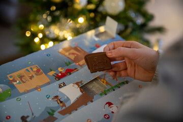 Fototapeta premium Gros plan d'un enfant ouvrant la fenêtre d'un calendrier de l'Avent avec un chocolat, sapin de noël avec guirlande lumineuse en fond.
