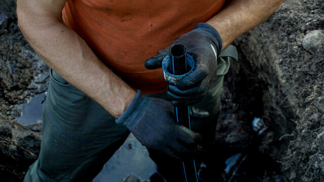 Worker installing plastic pipe connector onto irrigation line inside dug trench, highlighting repair process, water supply maintenance and hands on plumbing work in outdoor environment