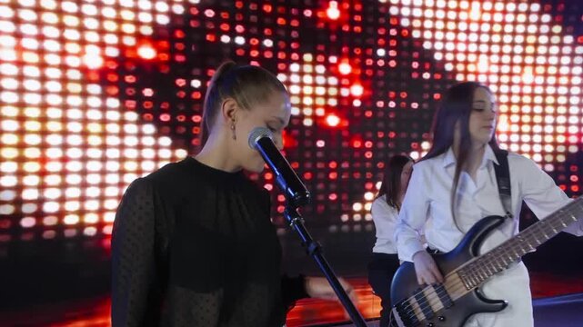 A young all female band performs on stage. A lead singer is at the microphone while a bassist plays her guitar. A drummer is in the background against a bright, colorful LED screen