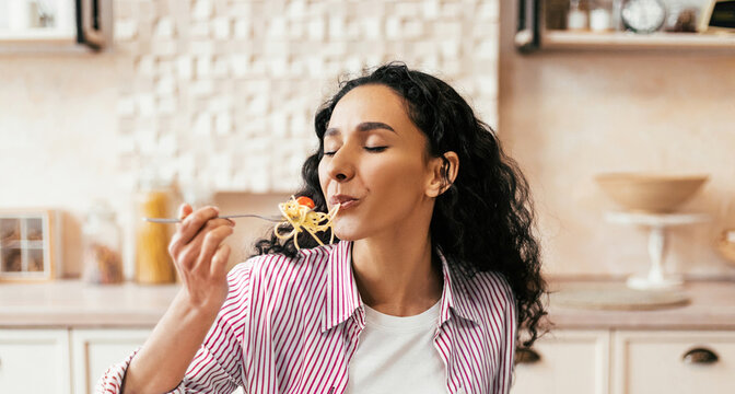 A woman is sitting at a kitchen table with a plate of pasta in front of her. She takes a bite, smiling as she enjoys her meal. The kitchen is light and inviting.