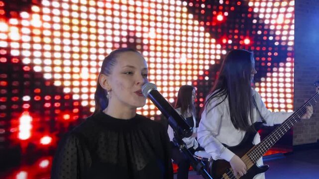 An all female music group performs on stage. A lead singer passionately sings into a microphone while a bassist plays next to her. A bright LED screen illuminates the background with red lights