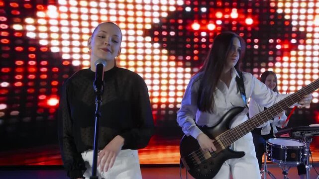 An all female music band performs on stage. The lead singer sings and plays keyboard while a guitarist plays bass. A bright LED screen with red lights is in the background. A modern live show