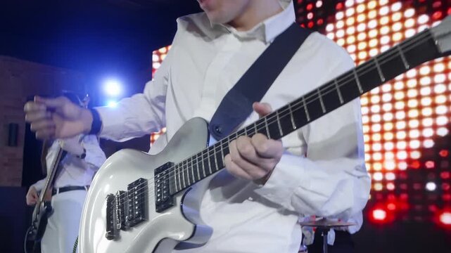 A young music band performs on a concert stage. A male guitarist in a white shirt plays a silver electric guitar, with a female bassist and drummer in the background against a bright LED screen