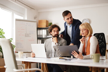 A business team works together in an office. They gather around a table focusing on a tablet while...