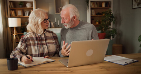 A couple sits at a table with a laptop open. They are discussing life insurance options. Notes and a coffee cup are nearby. The room has bookshelves and green plants. © Stockphotodirectors