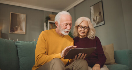 An elderly couple sits on a couch in their living room, looking at a tablet to explore life insurance options and discuss financial planning together during the evening hours. © Stockphotodirectors