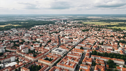 Obraz premium Aerial view of a densely populated city with orange roofs stretching towards green landscapes under a cloudy sky.