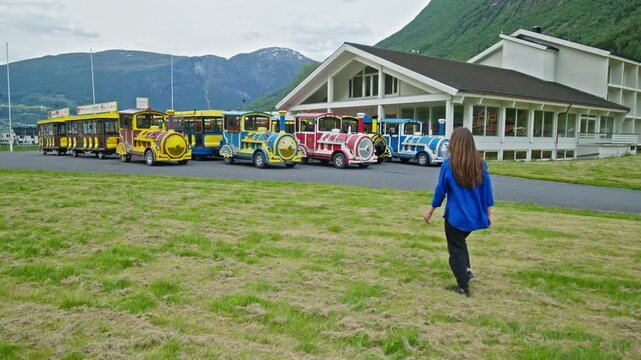 Tourist woman walks towards trains and modern building in Fl&aring;m. Scenic fjord village with mountains in Norway.