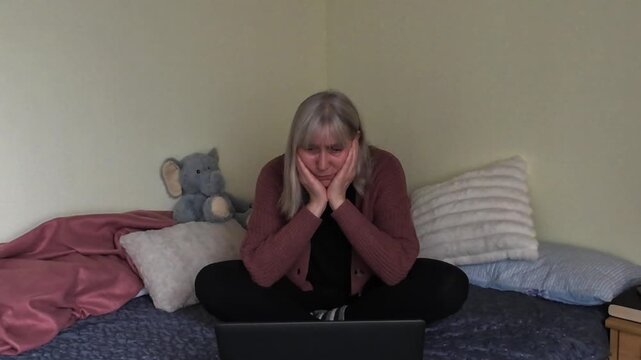 Mature woman feeling stressed while working on laptop at home sitting on bed