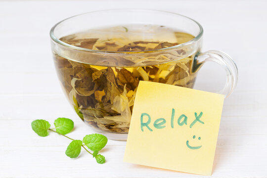 A glass cup of herbal tea with flowers, petals and leaves in it and yellow sticker note Relax on white wooden background macro