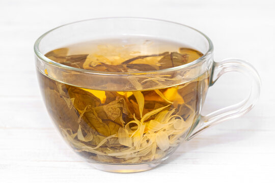 A glass cup of herbal tea with flowers, petals and leaves in it on white wooden background macro
