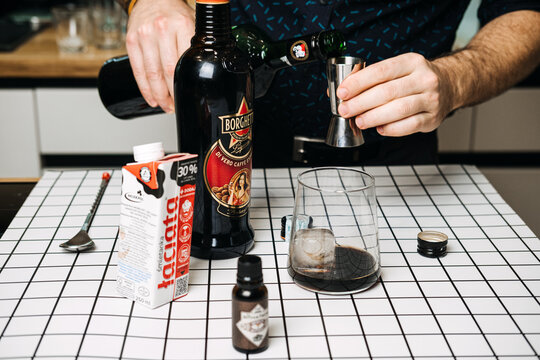 Person pours Cynar into jigger over grid surface with Borghetti liqueur and Taciata cream. At-home craft cocktail, amateur bartender skills, DIY specialty beverage. Warsaw, Poland, 20 November 2025