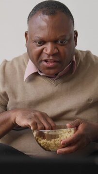 African american man watching TV and eating popcorn, vertical portrait at home in evening. Lazy weekend at home, senior single black male person enjoying favorite tv show or series, relax in apartment