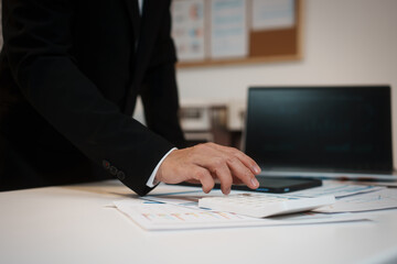 Fototapeta premium Businessman in suit using calculator on financial reports at office desk with laptop in background, analyzing budget and accounting data for corporate finance.