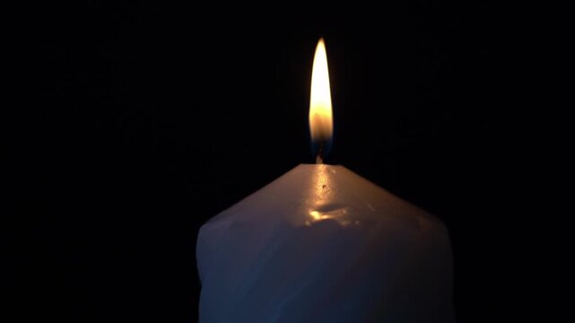 Single candle flame glows softly against black background. Candle wax melts gently around the wick's base. Light flickers, casting warm halo around the flame