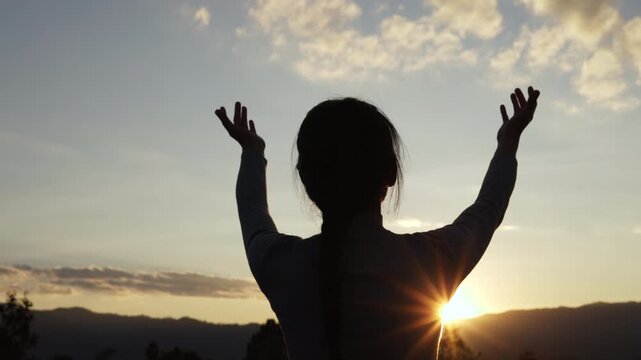 A silhouette of a woman open palm  praying on a mountaintop at sunset, 4K slow-motion video.